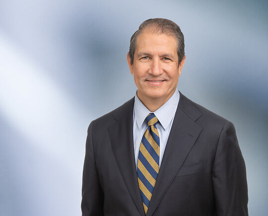 A man, Karl Butterer, in a dark checkered suit, white shirt, and blue striped tie is looking at the camera, standing against a blurred blue and white background.