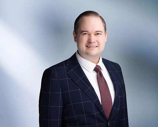 A man, Alexander Rusek, in a dark suit, light shirt, and patterned tie is smiling at the camera, standing against a blurred blue and white background.