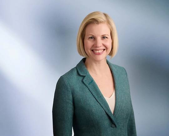 Woman, Mindi Johnson, with shoulder-length blonde hair wearing a black blazer, smiling against a blurred white and blue background.