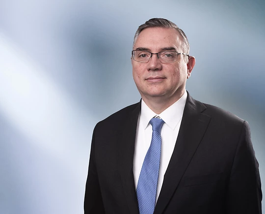A man, Clifford Hammond, in a dark suit, white shirt, and light blue tie is looking at the camera, standing against a blurred blue and white background.